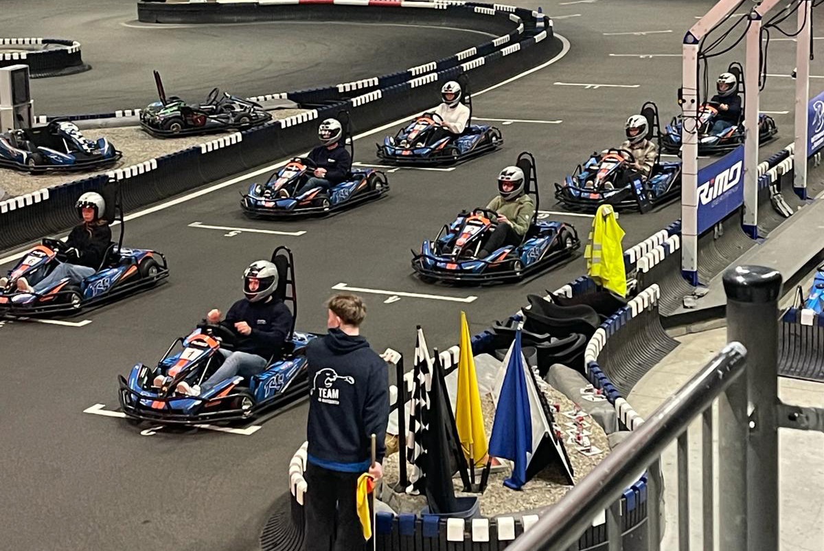 Mehrere Personen fahren in Karts über eine Indoor-Rennstrecke; ein Teammitglied steht am Streckenrand mit einer gelben Flagge.